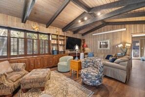 A spacious living room with a high ceiling, wooden beams, a large window with shutters, a fireplace, and a cozy seating area with patterned furniture. - Creekside Retreat | Fireplace + Gameroom (Rapid City)