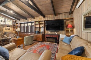 Un salon chaleureux avec un grand téléviseur à écran plat, des canapés en cuir, un tapis à motifs et des étagères en bois ornées de divers objets décoratifs.
