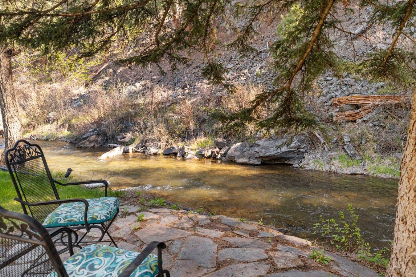 A stone patio with a chair and a table overlooking a clear stream, surrounded by trees and rocky terrain.