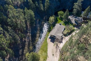 An aerial view of a house with a sloped roof, surrounded by dense forest and a river. - Creekside Retreat | Fireplace + Gameroom (Rapid City)