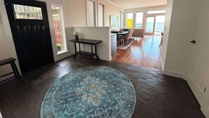 Un pasillo amplio con suelo de madera oscura, un tapete redondo con motivos y una zona de comedor con mesa y sillas, con vistas al exterior a través de grandes ventanales.