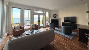 En stue med sofa, lænestole, pejs, fjernsyn og store vinduer med havudsigt.