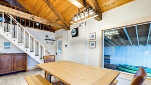 Une table à manger en bois avec des bancs, un escalier avec des rampes blanches et, par une grande fenêtre, une vue sur un bateau vert.