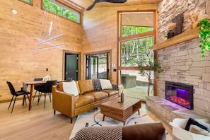 A cozy cabin interior with a stone fireplace, wooden walls, a leather sofa, and a dining area. - NEW Modern Cabin: Firepit, HotTub, Cozy Woods (Blue Ridge)