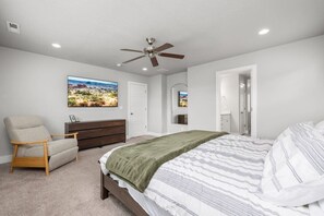 A bedroom with a bed, a chair, a dresser, a ceiling fan, and a wall-mounted TV.