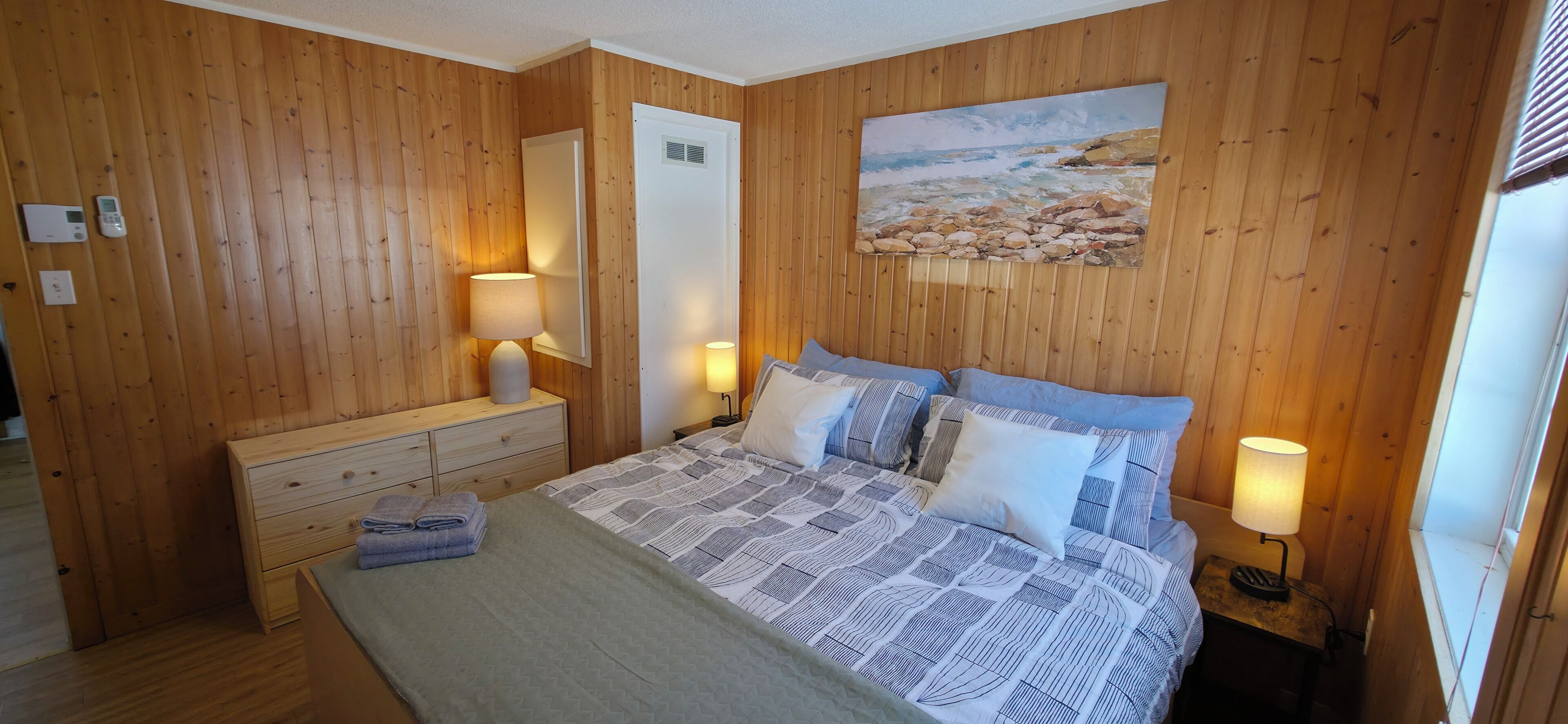 Une pièce aux murs lambrissés, comprenant un lit, des lampes de chevet, une commode et un tableau représentant une scène de plage.