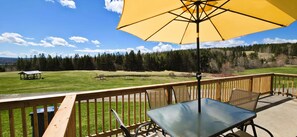 A deck with a table and chairs, a yellow umbrella, and a view of a grassy field and forest. - Spacious Cape Breton Getaway | King Bed | Perfect for Small Groups (Baddeck)
