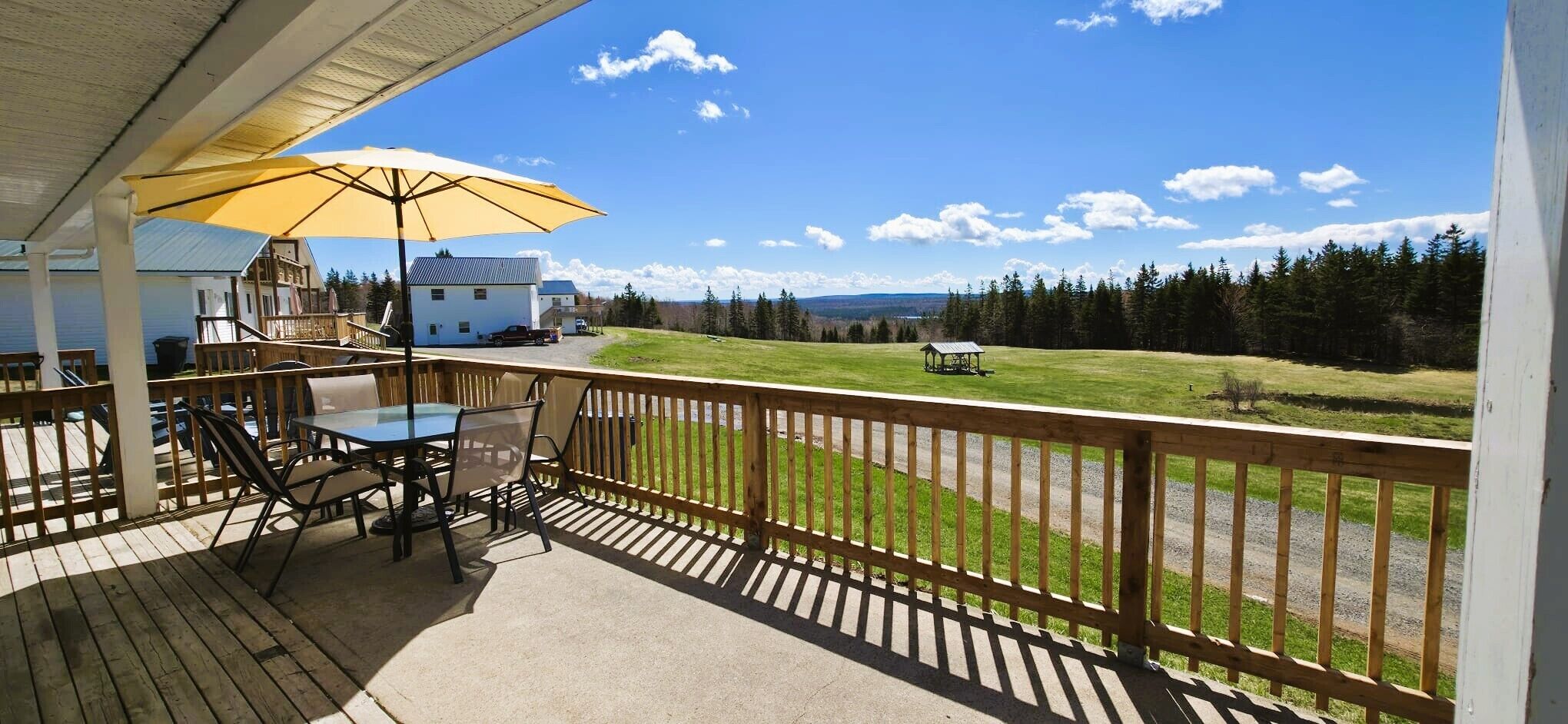 A covered outdoor area with a table and chairs, a yellow umbrella, and a view of a grassy field and trees.