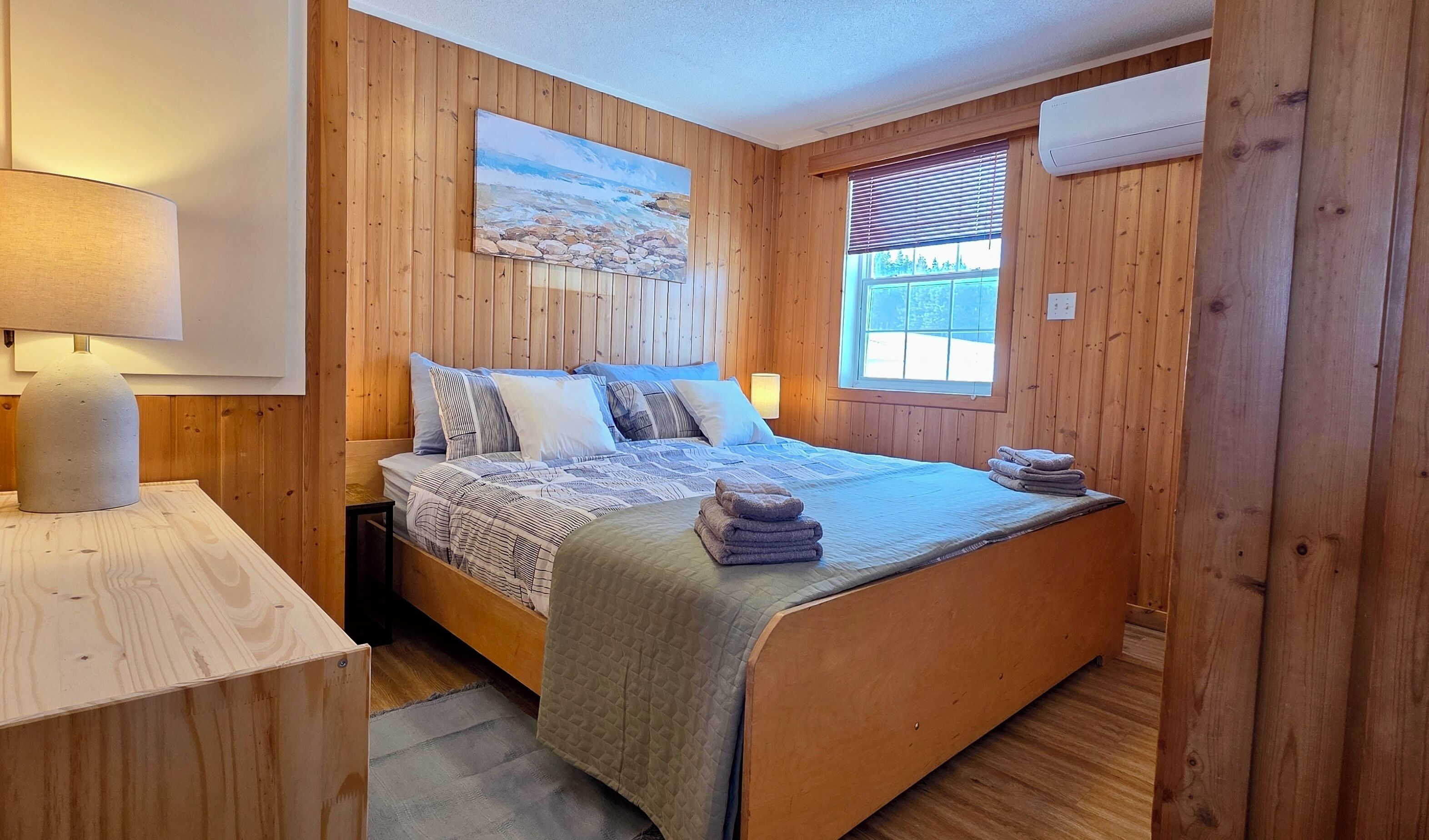 A wooden-paneled room with a bed, a painting, a window with blinds, and an air conditioning unit.