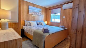 A wooden-paneled room with a bed, a painting, a window with blinds, and an air conditioning unit.