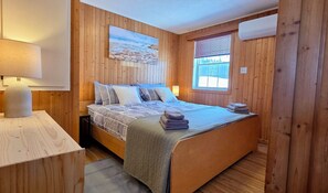A wooden-paneled room with a bed, a painting, a window with blinds, and an air conditioning unit. - Spacious Cape Breton Getaway | King Bed | Perfect for Small Groups (Baddeck)