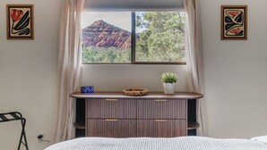Een slaapkamer met een bed, een nachtkastje met een klok, een raam met uitzicht op rode rotsformaties en twee kunstwerken aan de muur.