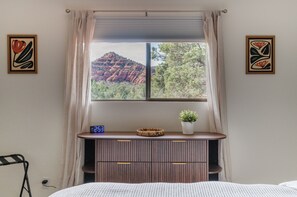 Ein Schlafzimmer mit einem Bett, einem Nachttisch mit einer Uhr, einem Fenster mit Blick auf rote Felsformationen und zwei an der Wand befestigte Kunstwerke.