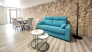 Quarto moderno com parede de pedra, sofá azul, mesa de jantar com cadeiras e uma mesinha redonda com um vaso.
