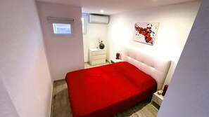 Ein Schlafzimmer mit roter Bettdecke, einem weißen Nachttisch, einem Beistelltisch, einer Wand-Klimaanlage, einem kleinen Fenster und einem abstrakten Kunstwerk an der Wand.