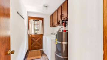 A compact kitchen with a washing machine, a stainless steel water heater, wooden cabinets, and a door with a brass handle.