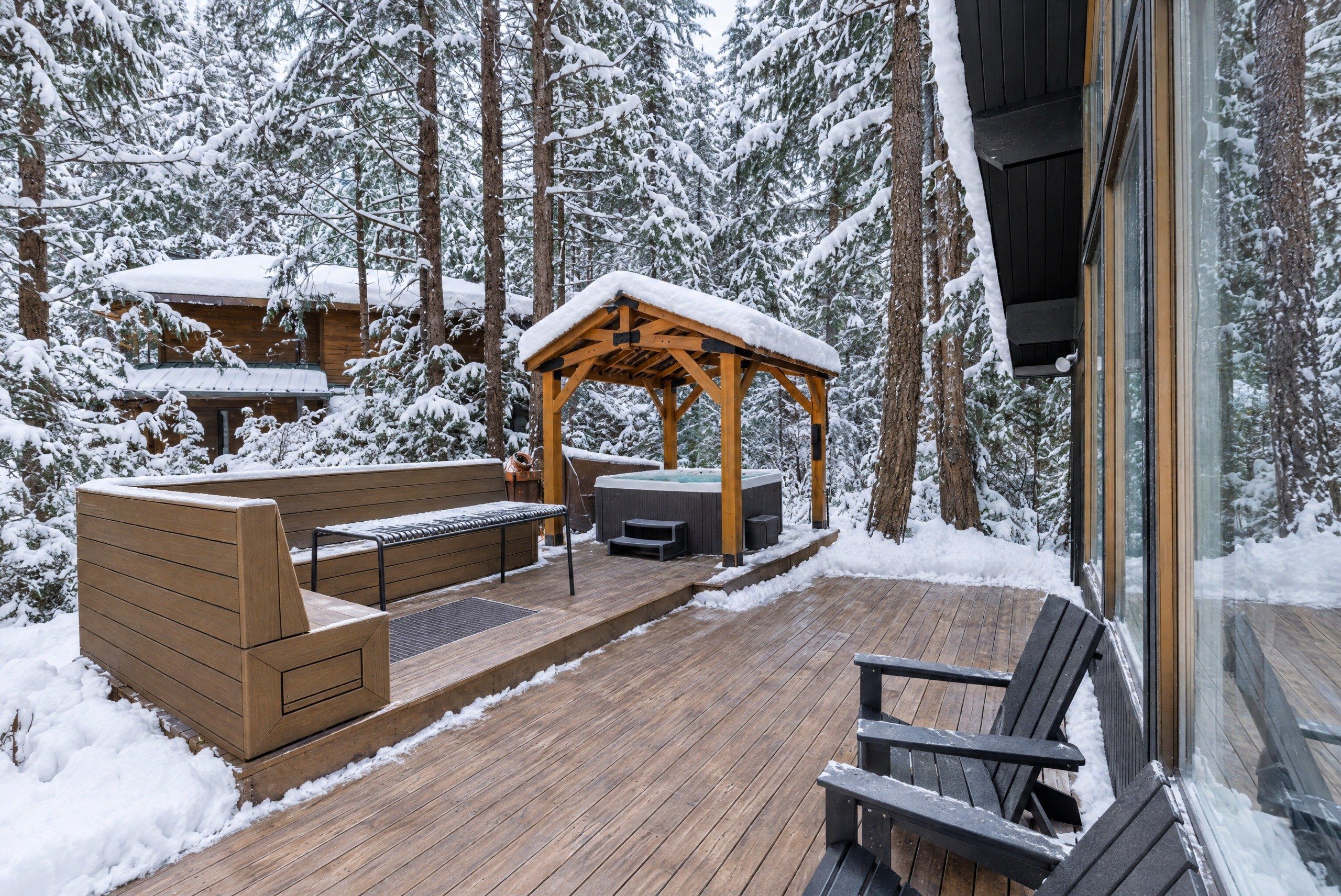 A wooden deck with a hot tub, seating area, and a covered gazebo in a snowy forest setting.