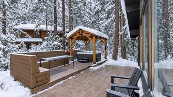 A wooden deck with a hot tub, seating area, and a covered gazebo in a snowy forest setting.