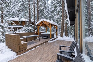 Een houten terras met een hottub, een zitgedeelte en een overdekte gazebo in een besneeuwd bos.
