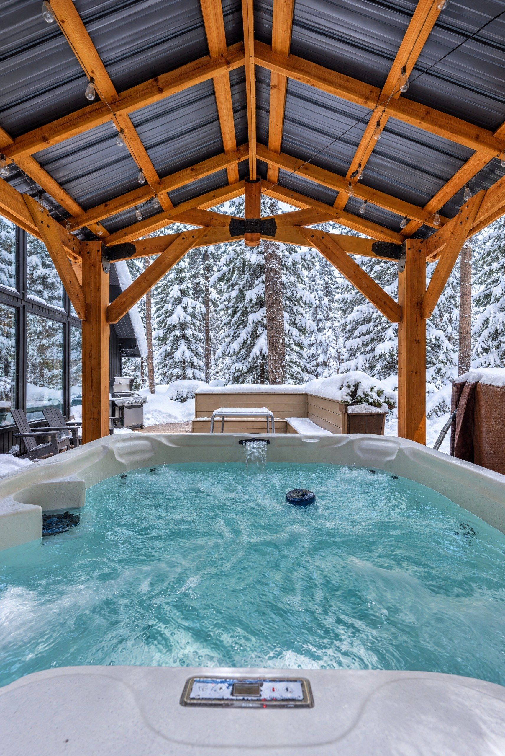 Un jacuzzi bajo una pérgola de madera con un bosque nevado al fondo.