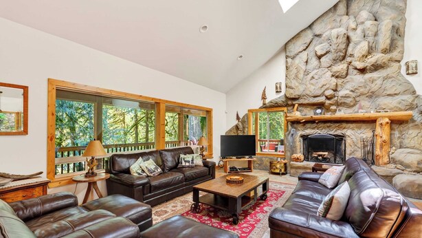 A spacious living room with leather sofas, a fireplace, and large windows overlooking a forest.