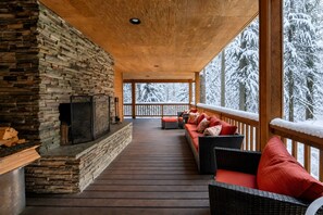 A covered porch with a stone fireplace, wooden deck, and a view of snow-covered trees.