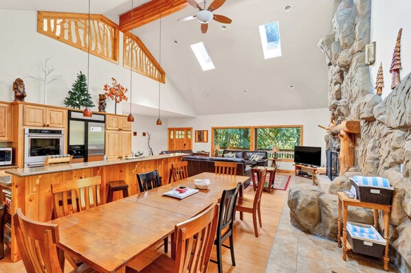 A spacious kitchen with wooden cabinets, a dining area with a wooden table and chairs, a stone accent wall, and a large window providing natural light. - 3 King Beds + Hot Tub + Game Room • Sleeps 12 (Rhododendron)