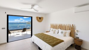 Quarto com cama, ventilador de teto, vista para o mar e varanda com cadeiras.