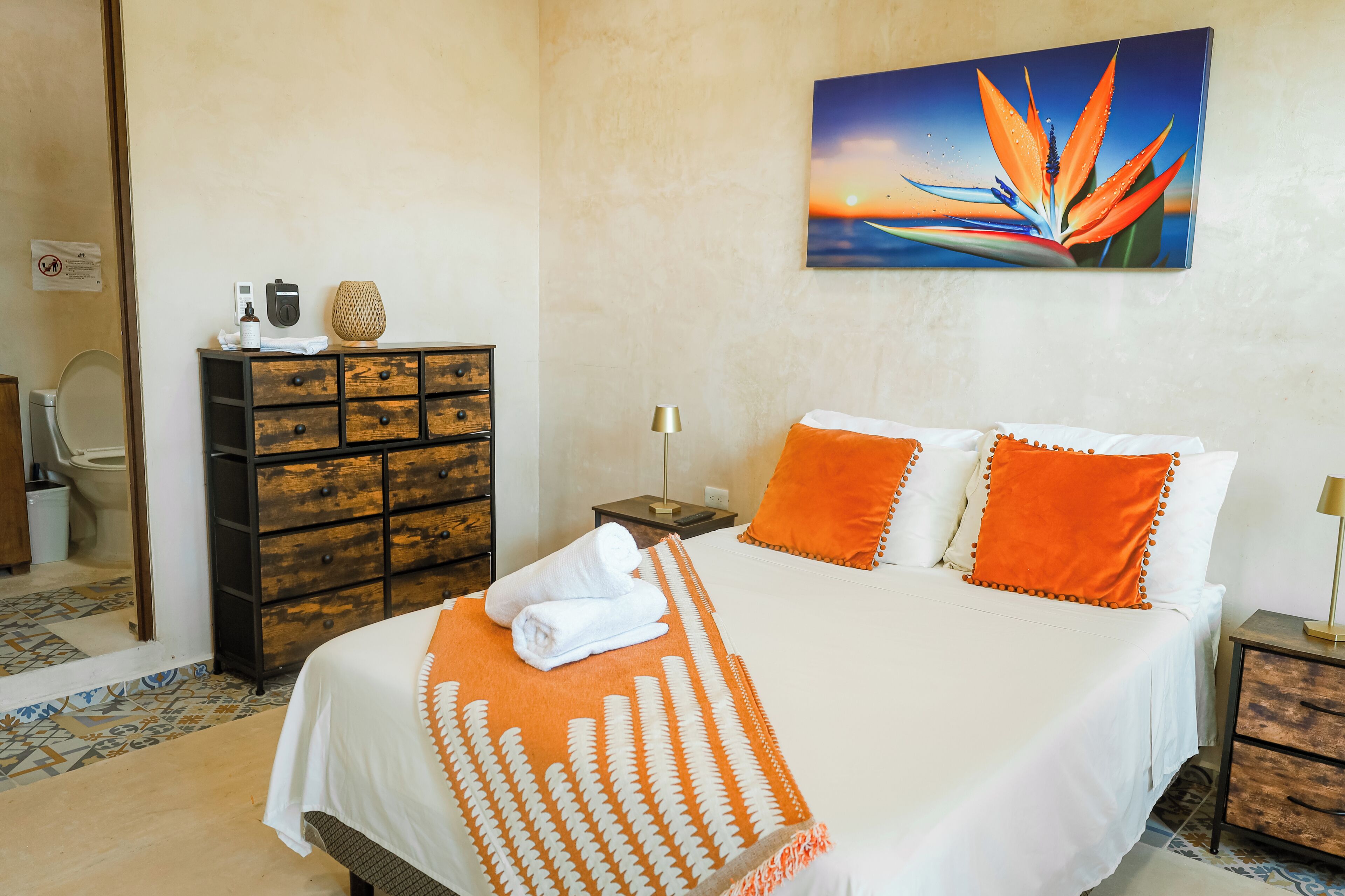 A neatly made bed with white and orange bedding, a wooden dresser with drawers, a bedside table with a lamp, and a wall-mounted painting of a bird of paradise.