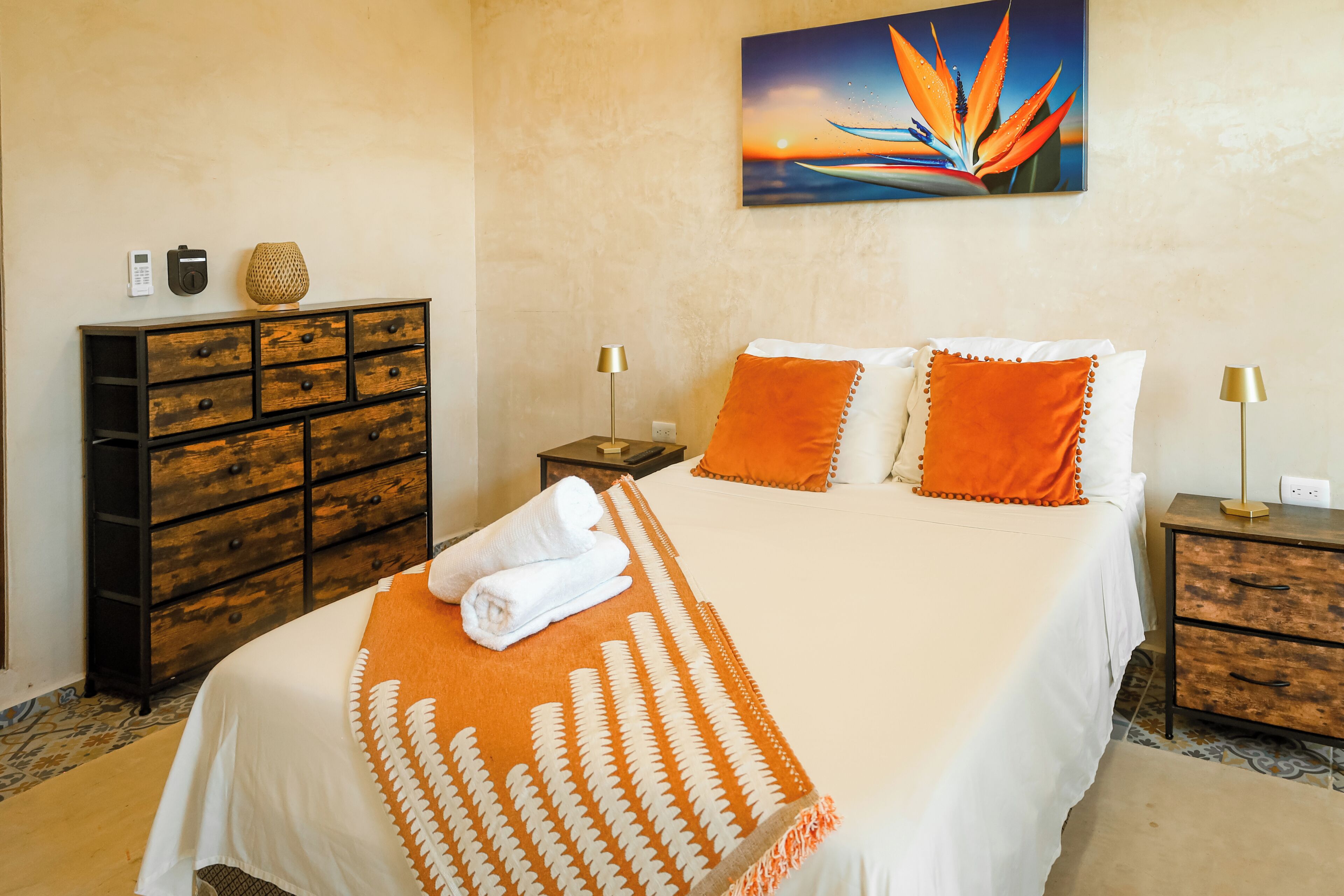 Quarto de hotel com cama, cómoda de madeira, mesa de cabeceira, abajur e um quadro na parede.