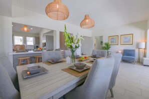 A modern living room with a dining area, grey upholstered chairs, a wooden dining table, and a kitchen in the background. - Mesa Vista - 4 BR Villa (Malmok)