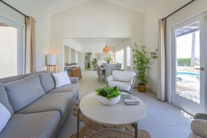 Ein geräumiges Wohnzimmer mit einer Couch, einem runden Couchtisch mit Pflanzen, einem Essbereich mit Stühlen und Blick auf einen Pool durch große Schiebetüren.