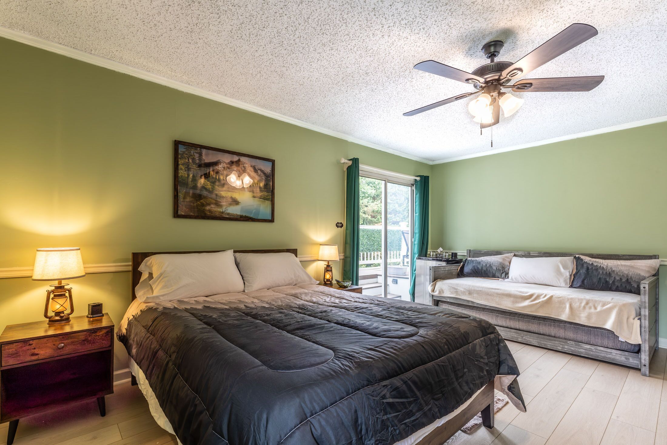 A bedroom with a bed, a sofa, a ceiling fan, a painting, and a nightstand with a lamp.