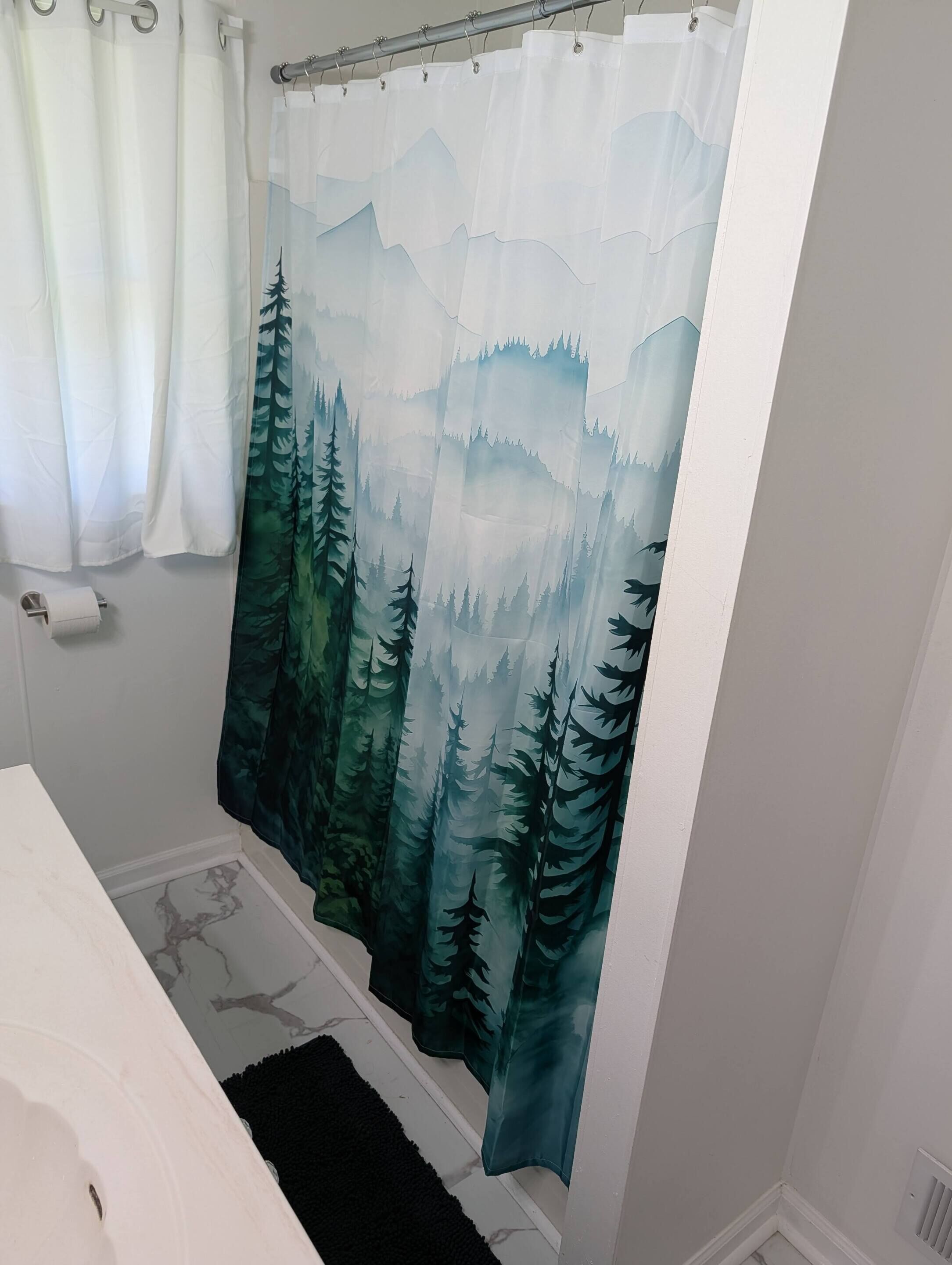 Un baño con cortina de ducha con motivos de bosque, un dispensador de papel higiénico y una alfombra negra.
