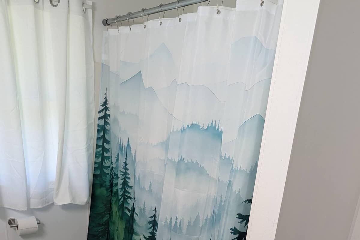 Un baño con cortina de ducha con motivos de bosque, un dispensador de papel higiénico y una alfombra negra.