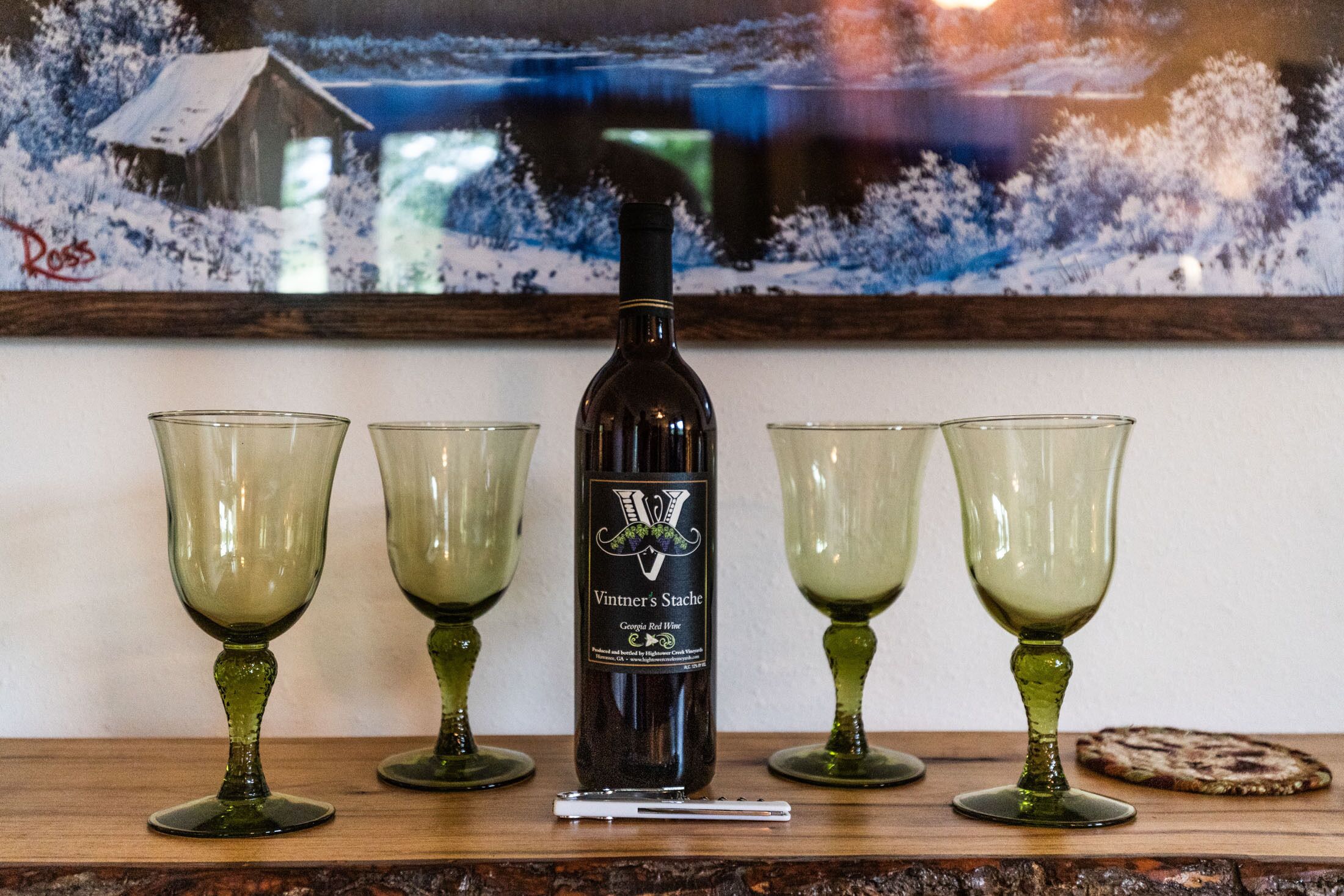 Une bouteille de vin rouge géorgien de la marque Vintner's Stache, accompagnée de quatre verres à vin verts, posée sur une surface en bois.