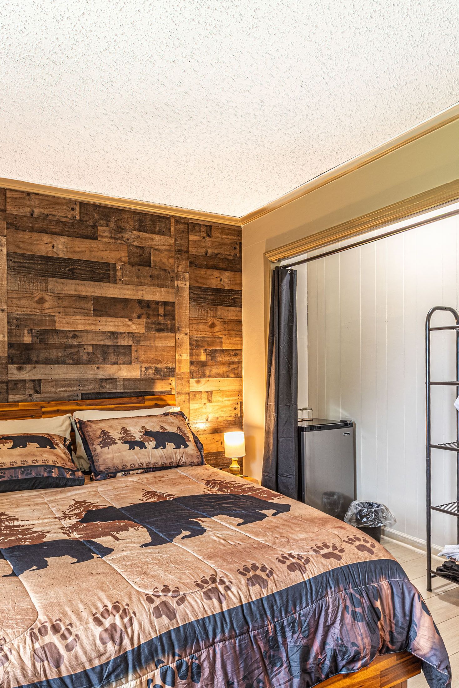 A bedroom with a wooden headboard, a bed with a bear-themed duvet, a nightstand with a lamp, a mini fridge, and a metal rack.