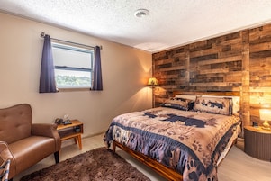 Un dormitorio con cabecera de madera, una cama con colcha estampada, un sillón de cuero, una mesita de noche de madera y una ventana con cortinas.