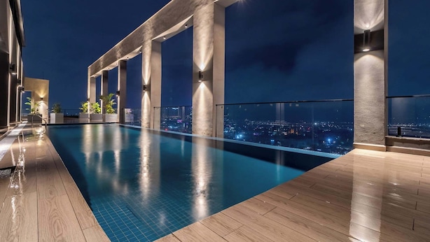 Ein Pool mit unendlicher Optik auf einem Dach, mit Blick auf die Stadt bei Nacht, gestützt von Betonstützen und mit Holzbelag.
