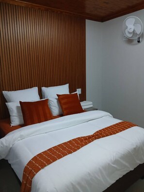 Ein Bett mit weißer und orangefarbener Bettwäsche, einem Holzschrankgitter-Kopfstück und einem an der Wand befestigten Ventilator.