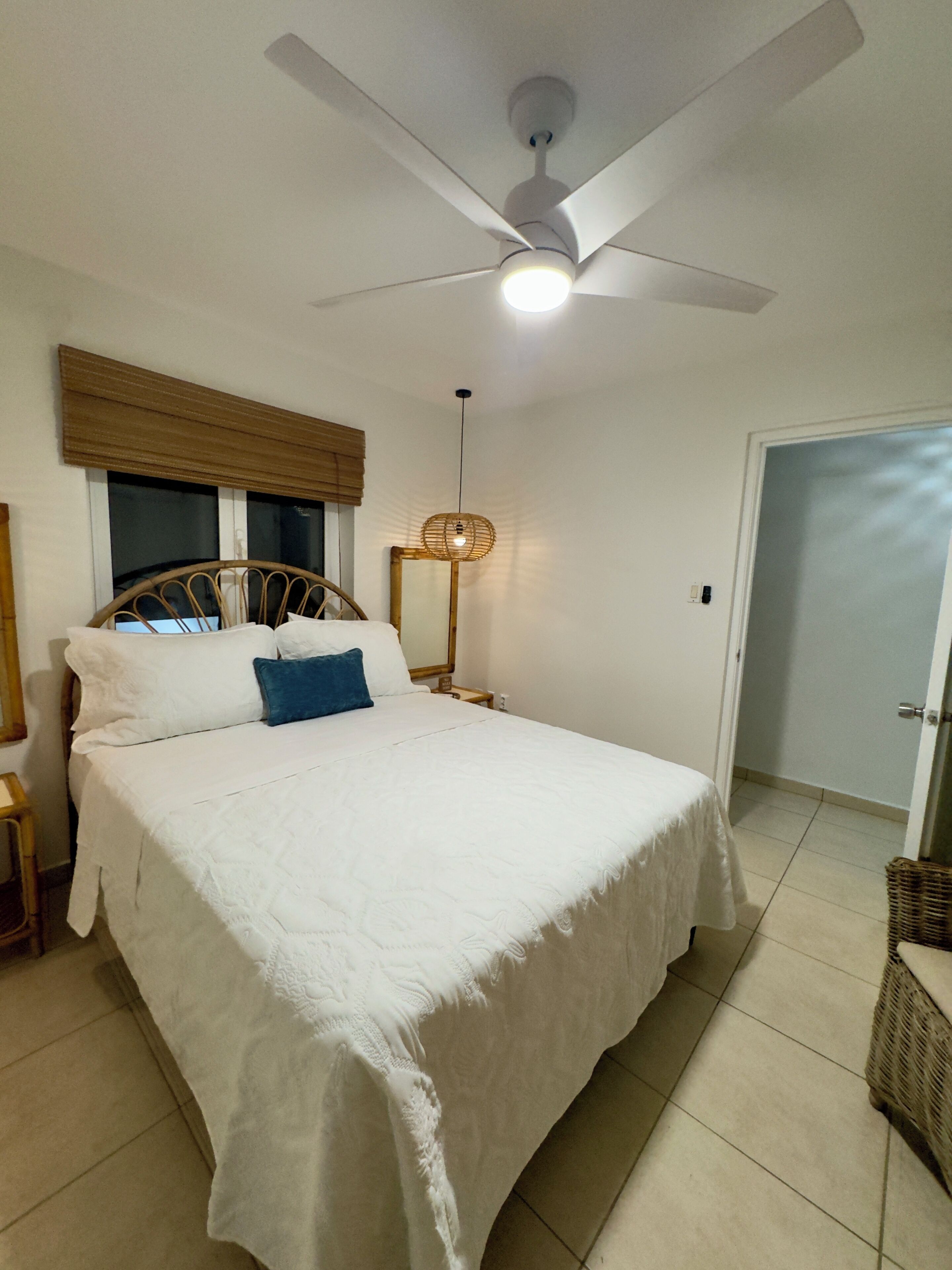 A bedroom with a bed, a ceiling fan, a bedside lamp, a window with blinds, and a door.