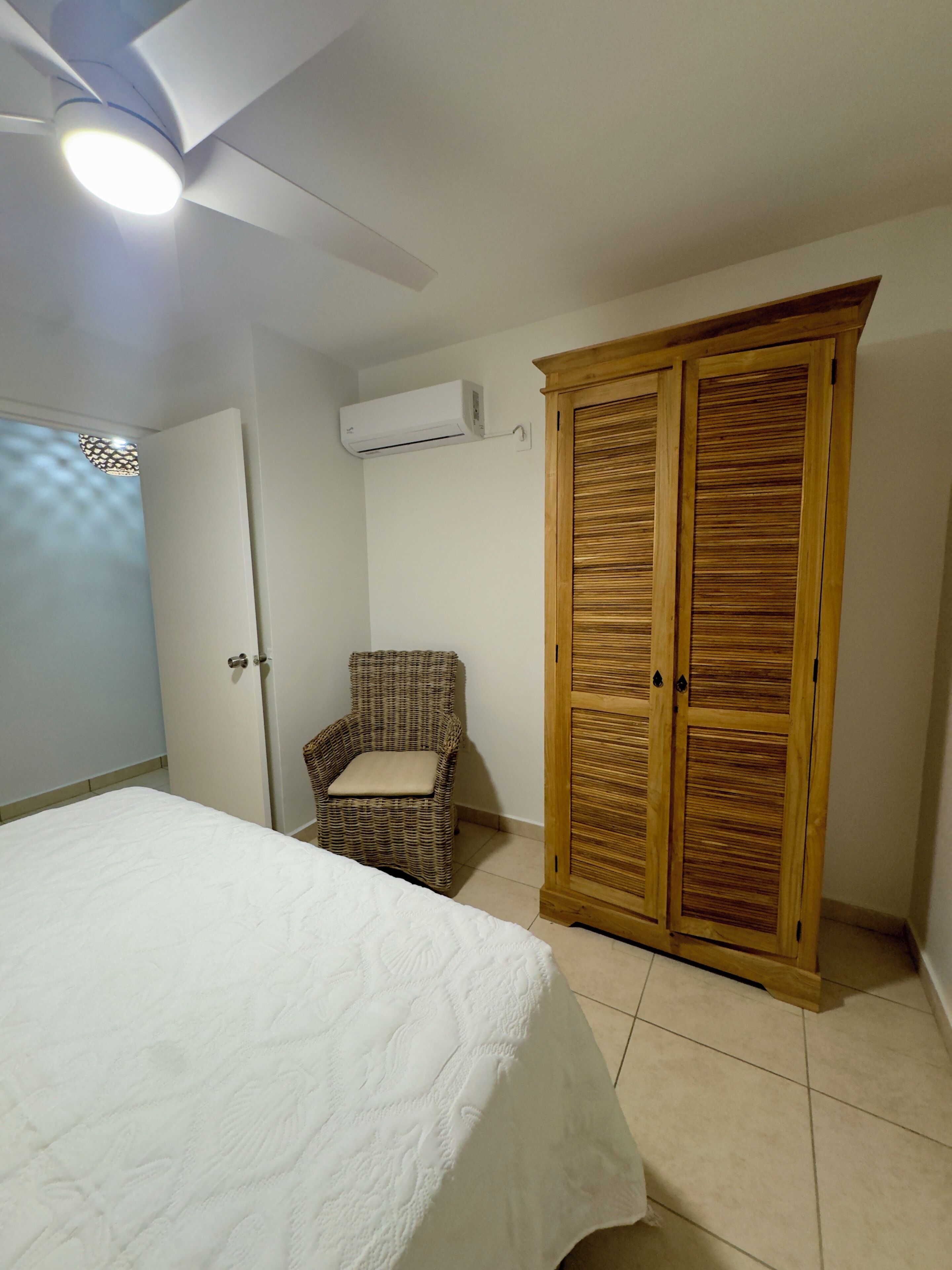 Une chambre à coucher comprenant un lit, une chaise en osier, une armoire et un appareil de climatisation.