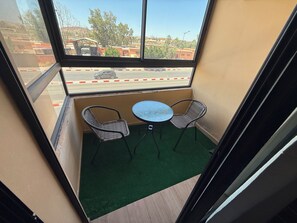 Ein kleiner Balkon mit zwei Korbstühlen und einem runden Tisch, mit Blick auf eine Straße mit Häusern und Bäumen.