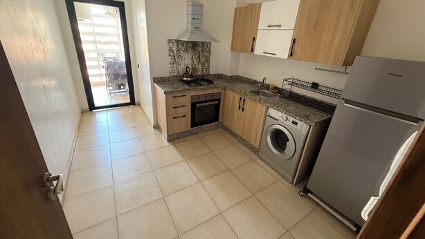 A compact kitchen with a washing machine, a refrigerator, and wooden cabinets. - Nour 5 Modern Apartment with Pool & Parking Near Train Station Central Marrakech (Marrakesh)