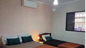 Une chambre avec deux lits, un ventilateur de plafond, un appareil de climatisation et une fenêtre munie d’une grille métallique.