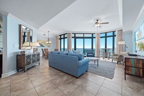 Un salón espacioso con un sofá azul, un comedor con sillas, un ventilador de techo y grandes ventanales con vistas.