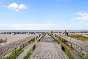 Ein hölzerner Steg führt zu einem Strand mit blauen Sonnenschirmen und Liegestühlen.