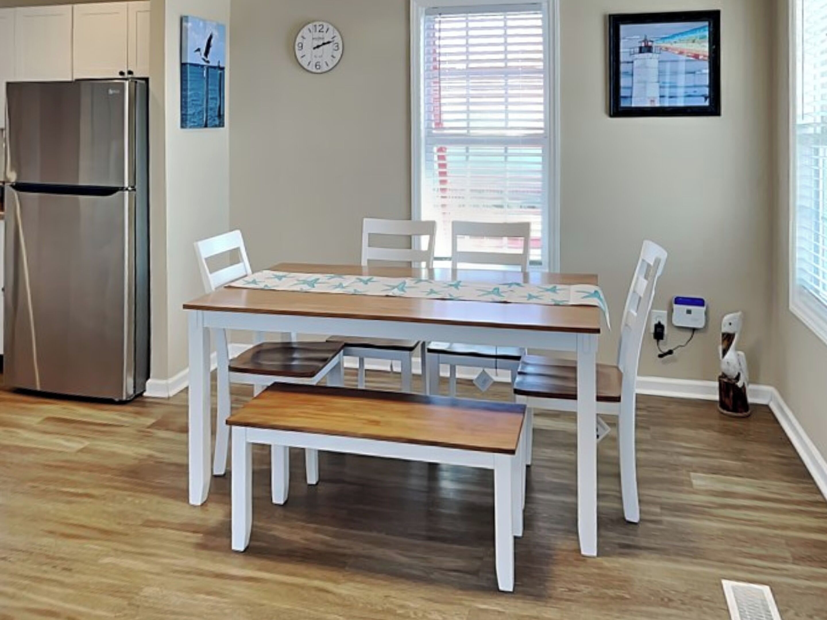 Un coin repas avec une table en bois, des chaises blanches et un banc. On aperçoit un réfrigérateur en acier inoxydable à l’arrière-plan.