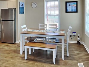 Comedor con mesa de madera, sillas blancas, una banca, refrigerador de acero inoxidable, un reloj de pared y una ventana con persianas.