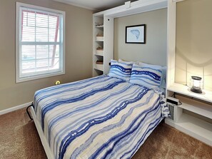 Un dormitorio con cama, ventana con persianas, estante con adornos y un cuadro enmarcado en la pared.